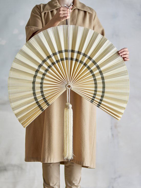 Persoon in beige jas houdt een grote beige waaier met zwarte strepen en een kwast vast.