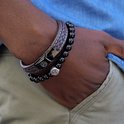 Close-up van een bruine gevlochten leren armband met witte stiksels en een zwarte sluiting met leeuwenlogo, gedragen naast een zwarte kralenarmband.