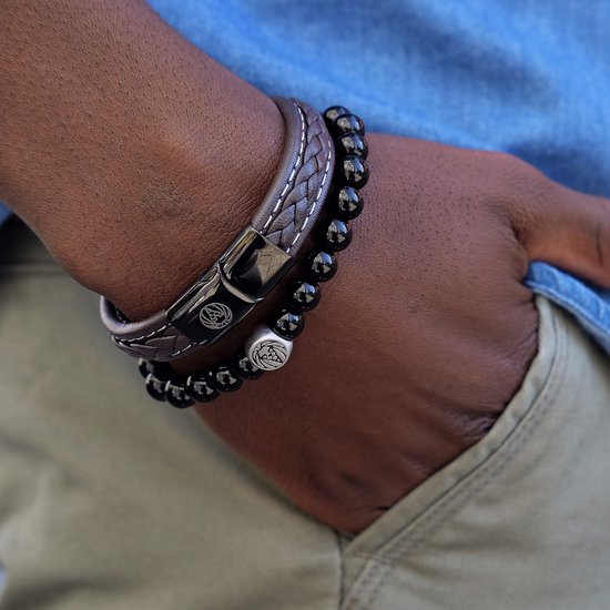Close-up van een bruine gevlochten leren armband met witte stiksels en een zwarte sluiting met leeuwenlogo, gedragen naast een zwarte kralenarmband.
