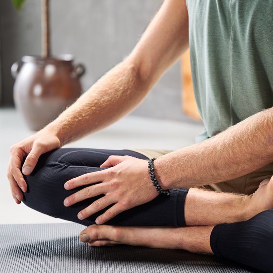 Man zit in kleermakerszit op een mat, draagt een groen tanktop en een dubbele rij armband met zwarte en grijze kralen.