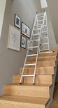 Aluminium trapadapterladder op een houten trap, met ingelijste kunst aan de muur.