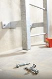 Drie metalen pluggen met moeren en ringen op een betonnen vloer, naast een metalen frame gemonteerd aan de muur.