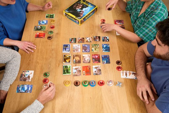Vier mensen spelen het bordspel Splendor aan een houten tafel, met kaarten en fiches.