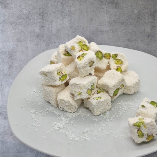 Vierkante stukjes wit Turks fruit met pistachenoten en kokos. Gestapeld op een lichtgrijs bord met een grijze achtergrond.