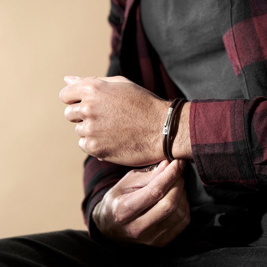 Detailopname van een man die een bruine leren armband met zilverkleurige sluiting aan zijn pols vastmaakt.