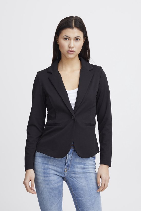 Vrouw met lang donker haar draagt een zwarte blazer en spijkerbroek. Witte top.