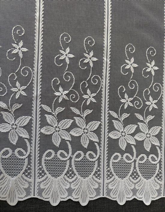 Detail van een wit kantengordijn met bloemen, bladeren en een decoratieve rand.