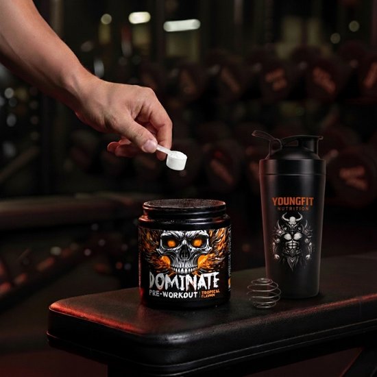 Dominate pre-workout tropische smaak, zwarte pot met schedel-afbeelding. Naast een zwarte shaker met YoungFit Nutrition logo. Vinger voegt schepje toe.