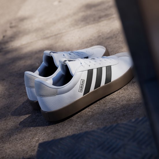 Twee witte leren Adidas VL Court 3.0 sneakers met bruine zool, zwarte strepen en Adidas logo, op een betonnen ondergrond.