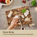 Une planche à découper Namture en bois L, sur laquelle des champignons et du persil sont coupés. Une main coupe avec un couteau.