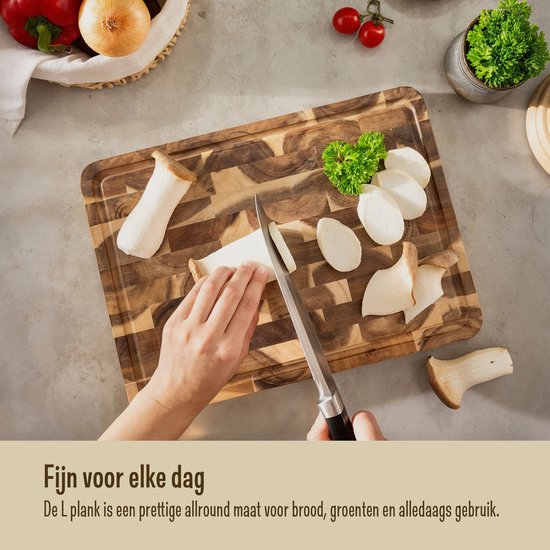 Une planche à découper Namture en bois L, sur laquelle des champignons et du persil sont coupés. Une main coupe avec un couteau.