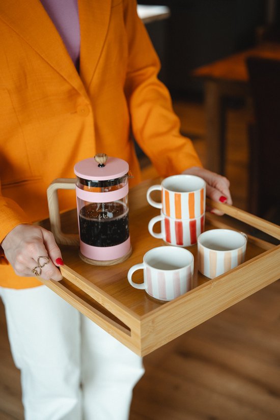 Vrouw in oranje jas houdt een houten dienblad met een roze koffiepot en vier gestreepte kopjes vast.