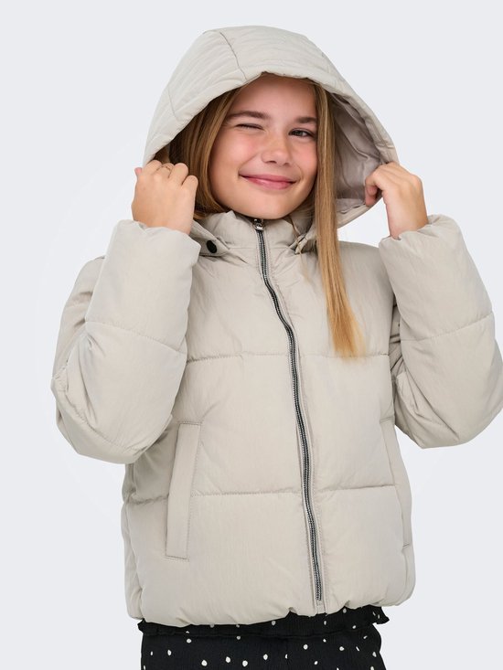 Meisje met blonde staartjes, draagt een lichtbeige pufferjack met capuchon, knipoogt en trekt aan de capuchon.