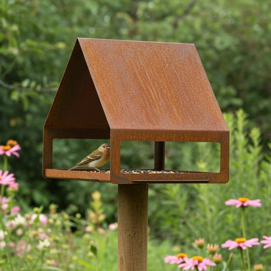 Roestkleurig vogelvoederhuis met puntdak op een paal, in een bloementuin. Een vinkje zit op de rand.