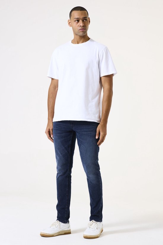Man draagt een effen witte T-shirt en een donkerblauwe slim fit jeans met een lichte wassing.