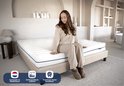 Een vrouw in een beige pyjama zit op een wit matras met horizontale lijnen op een beige bedframe. Vlag van Nederland en teksten over orthopedisch matras en hotelkwaliteit.