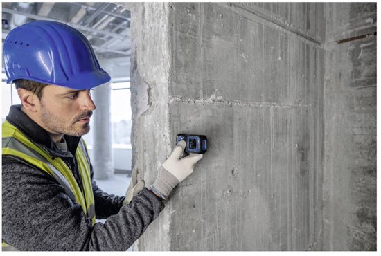 Man met blauwe helm en gele handschoenen houdt laserafstandsmeter vast tegen betonnen muur.