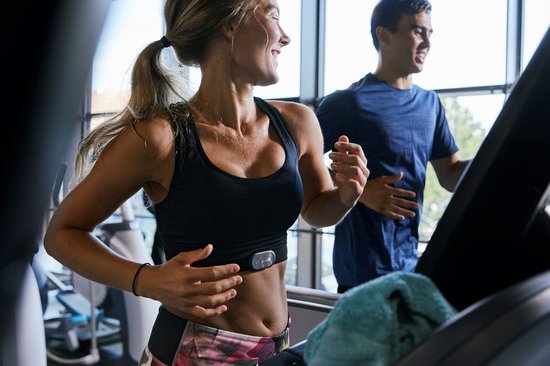 Vrouw met paars-roze sportbroek en zwart sportshirt draagt zwarte Polar hartslagsensor op loopband.