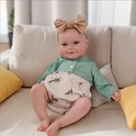 Baby met een beige strik, groen shirt en luierbroekje met olijvenprint, zittend op een bank.