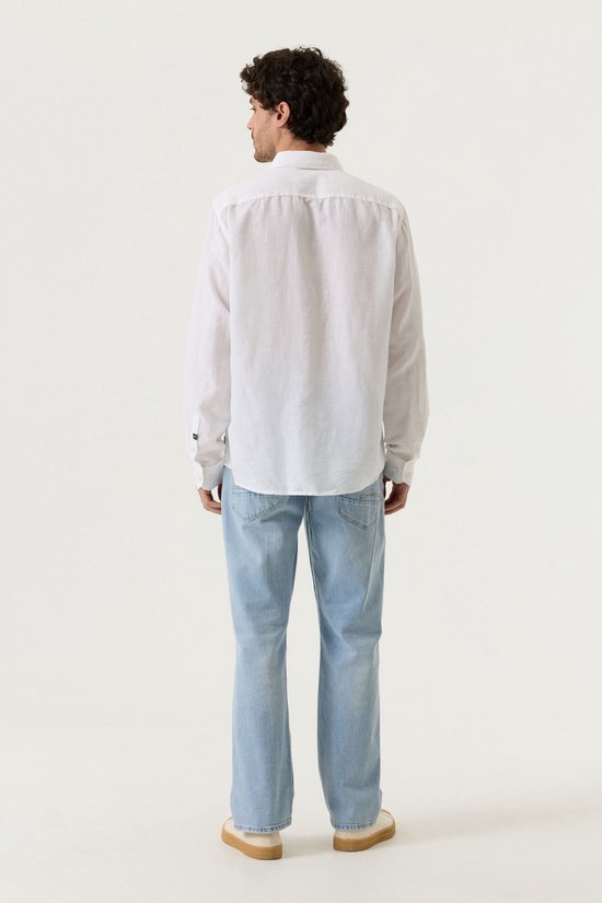 Man met krullend haar, van achteren gezien, draagt een wit linnen overhemd met lange mouwen en lichtblauwe jeans.