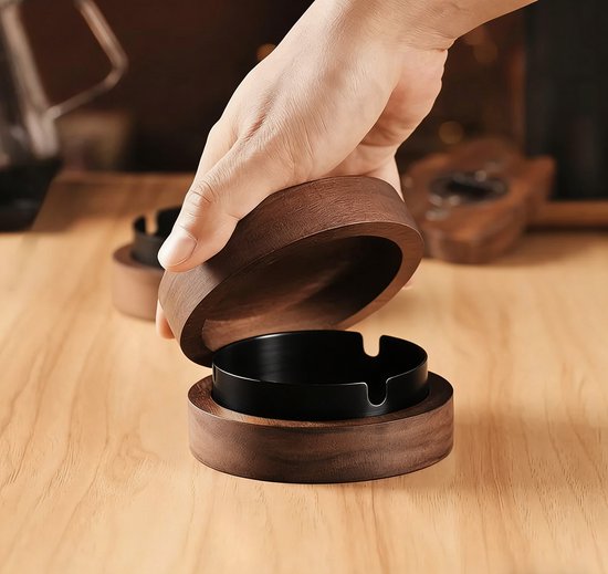 Een ronde houten asbak met een zwart metalen inzetstuk. Een hand opent het deksel, dat ook van hout is.