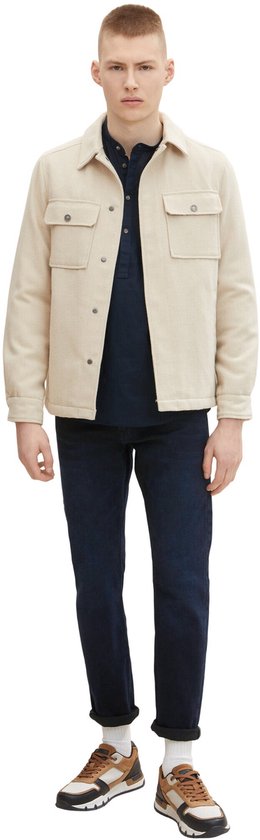 Man in een donkerblauwe jeans, donkerblauw poloshirt en een crèmekleurig jack.