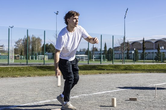 Jonge man in een wit T-shirt en zwarte broek gooit met een Kubb-pion op een sportveld.