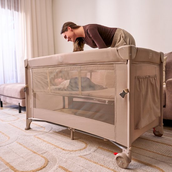 Lichtbruin reisbedje met gaaszijde, een baby slaapt binnenin. Een vrouw kijkt naar binnen.