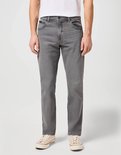 Man staat vol in beeld in een grijze jeans en een wit t-shirt. Hij draagt lichte sneakers.