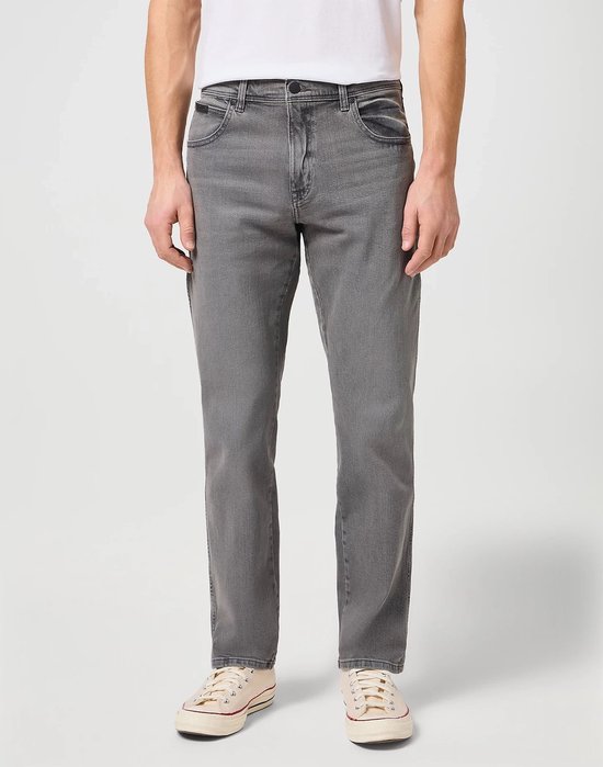 Man staat vol in beeld in een grijze jeans en een wit t-shirt. Hij draagt lichte sneakers.
