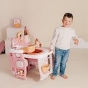 Jongen met witte trui en spijkerbroek lacht naar camera bij roze poppenverzorger.