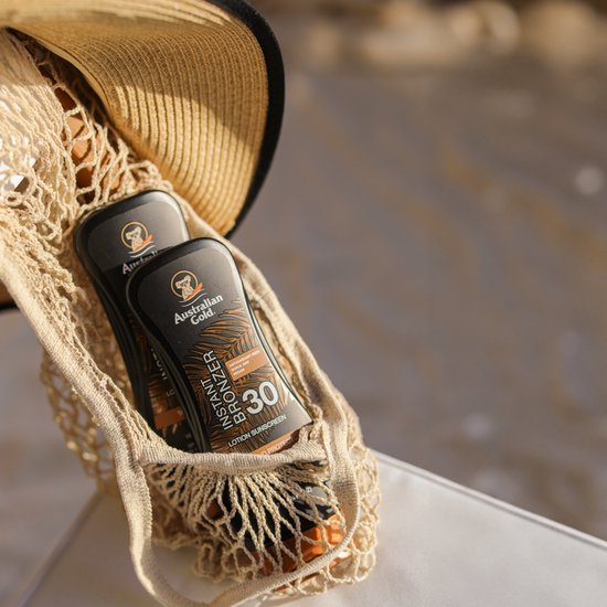 Twee flessen Australian Gold Instant Bronzer SPF 30 in een rieten tas met een strooien hoed.