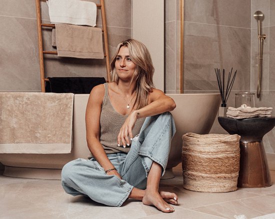 Een vrouw met blonde haren zit op de grond in een badkamer, met beige handdoeken op een houten rek op de achtergrond.