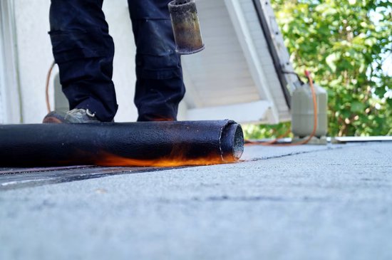 Man in werkbroek gebruikt brander op zwarte dakrol, met vlammen.