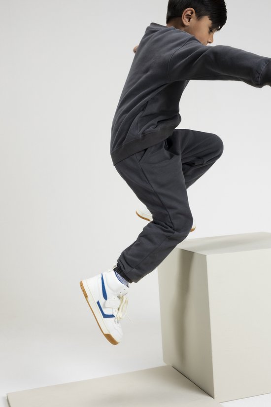 Jongetje in donkergrijs joggingpak, springend met witte sneakers met blauwe accenten.
