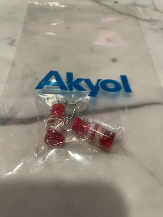 Gouden oorbelletjes in de vorm van rode flesjes met witte inhoud, verpakt in plastic.