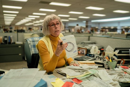 Een oudere vrouw met bril en grijs haar zit aan een bureau vol papieren en houdt een document met 'PAID' erop omhoog.