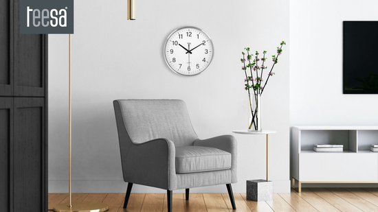 Een zilverkleurige wandklok met witte wijzerplaat hangt aan een witte muur boven een grijze fauteuil.