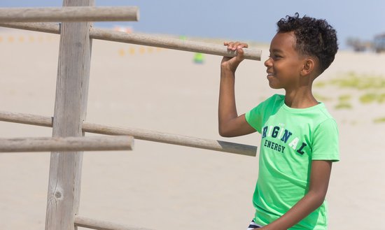 Jongetje met krullend haar in een felgroen T-shirt met de tekst 'ORIGINAL ENERGY' en een houten klimrek op het strand.
