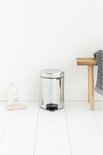 Briljant stalen Brabantia NewIcon prullenbak met zwart voetpedaal, naast een houten bankje met handdoek.