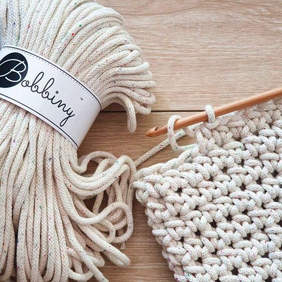 Roomwitte Bobbiny macramé garen met gekleurde spikkels, naast een haakproject.