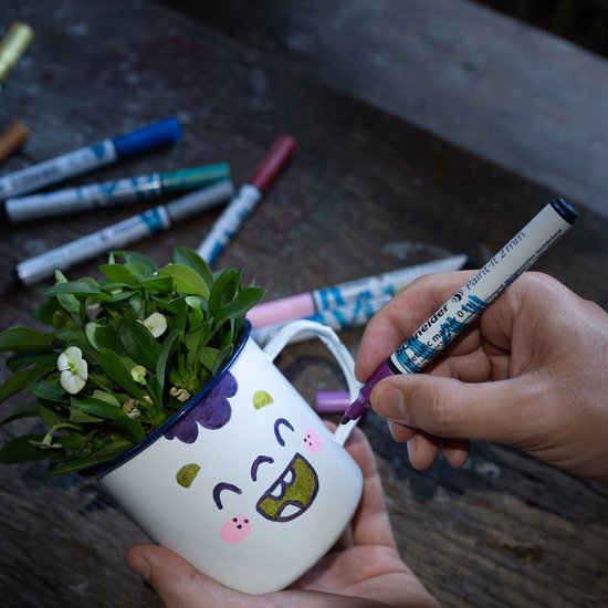 Hand met een paarse metallic stift tekent op een wit mokje met gezichtje. Mokje bevat plantjes.