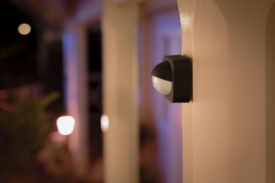 Zwarte Philips Hue buitenbewegingssensor bevestigd aan een witte zuil, met gedimde lichten op de achtergrond.