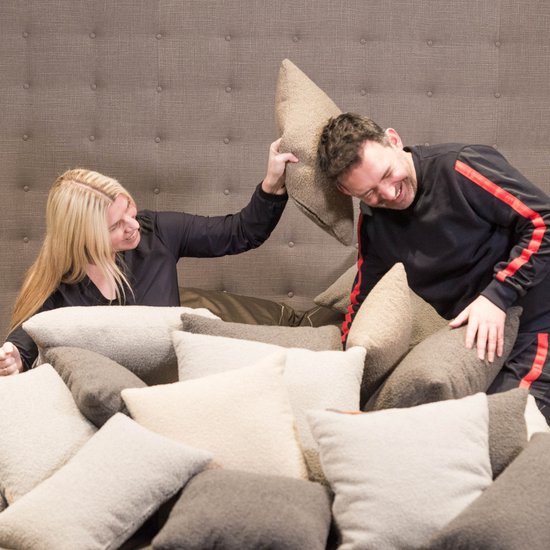 Een vrouw met blond haar gooit een kussen naar een lachende man in een donkere woonkamer vol kussens.