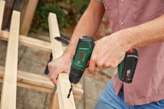 Man met gestreepte blouse boort in een houten constructie met een groene Bosch boormachine.