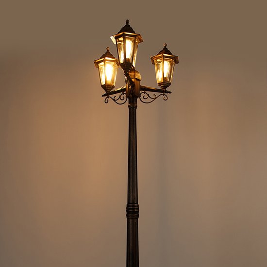 Een drielichts lantaarnpaal in de kleur goud/messing met verlichte lampen, tegen een donkere achtergrond.