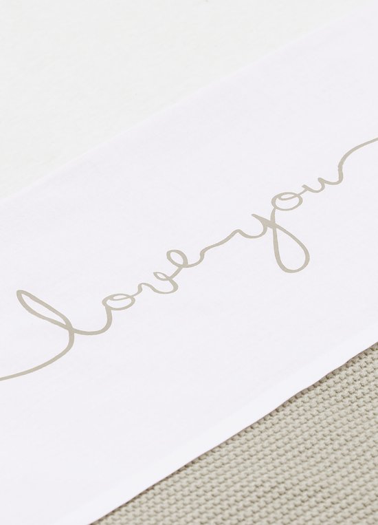 Detail van een wit ledikantlaken met 'love you' tekst, deels over een crème gebreid laken.