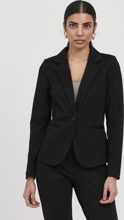 Vrouw met donker haar draagt een zwarte blazer en broek. Gouden oorbellen.