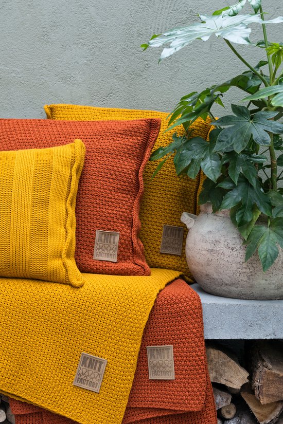 Gele en oranje gebreide kussens en plaid naast een plant.