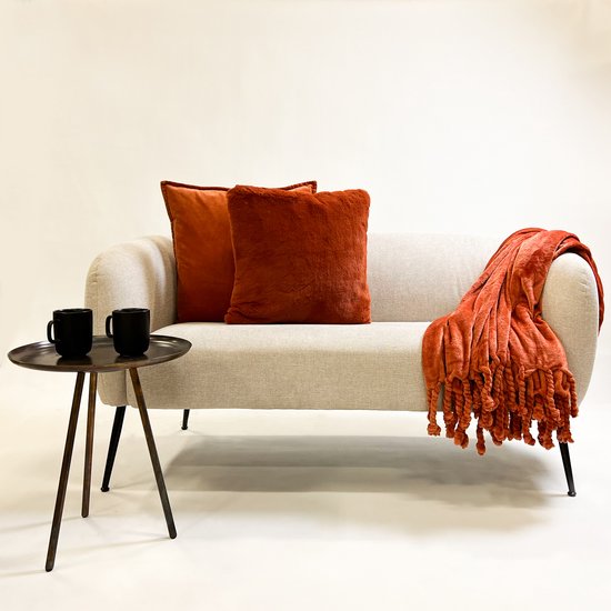 Grijze bank met twee oranje kussens (één velours, één fluffy) en een oranje plaid met flosjes.